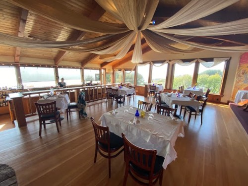 A spacious restaurant interior with wooden beams, tables set for dining, and large windows offering a scenic view.