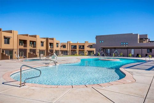 A clear blue pool surrounded by modern buildings under a bright blue sky. Sun loungers are placed around the pool area.