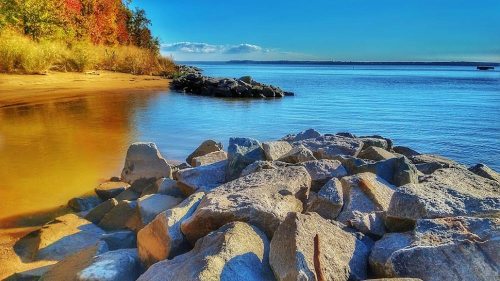 A serene lakeside view with colorful trees, smooth rocks, and calm water under a clear blue sky.