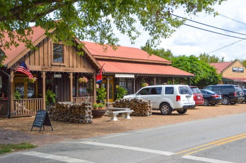 A rustic wooden building with a red roof, surrounded by parked cars and greenery, featuring flags and a sign that says "OPEN."