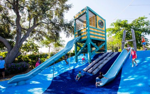 A colorful playground with two slides, a climbing structure, and children playing on blue rubber surfacing.