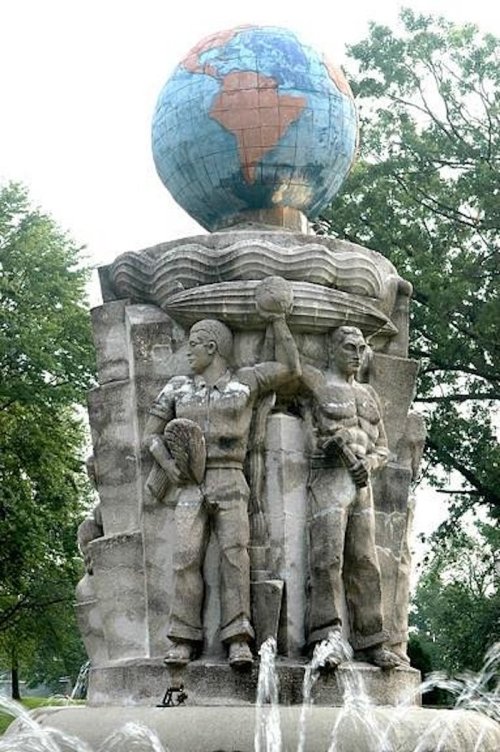 A fountain sculpture featuring two muscular figures holding a globe, surrounded by flowing water and greenery.