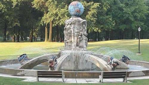 A decorative fountain featuring a globe atop a sculpted base, surrounded by benches and lush green trees.