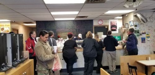 A busy restaurant scene with customers waiting at the counter and staff preparing food in a casual setting.