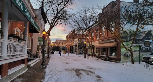 Snow-covered street lined with charming buildings, festive lights, and a mural, under a colorful sunset sky.