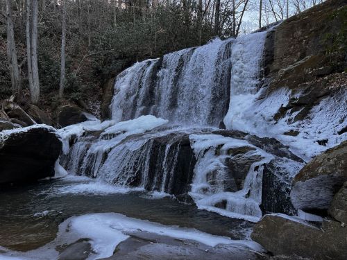 A frozen waterfall cascades over rocky ledges, surrounded by trees and a serene winter landscape.