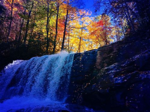A vibrant waterfall cascades over rocks, surrounded by colorful autumn foliage and a clear blue sky.