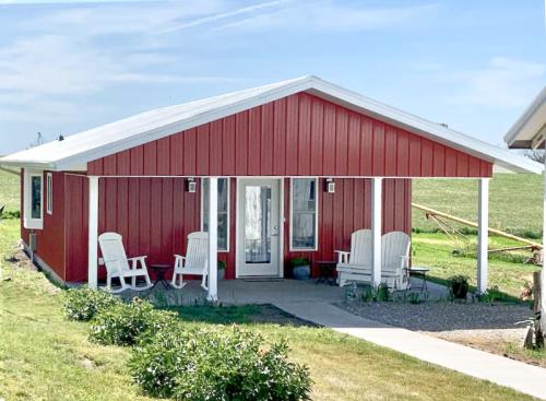 A red cottage with a white roof, featuring two chairs on the porch and a grassy field in the background.