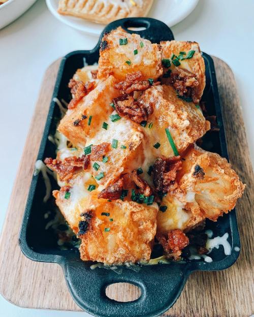A skillet of crispy, golden tater tots topped with melted cheese and chopped bacon, garnished with green chives.