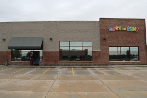 Exterior of a brick building with large windows and a colorful sign reading "LOST IN FUN!" on a cloudy day.