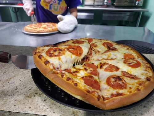 A freshly baked pepperoni pizza on a black tray, with a second pizza being placed in the background.