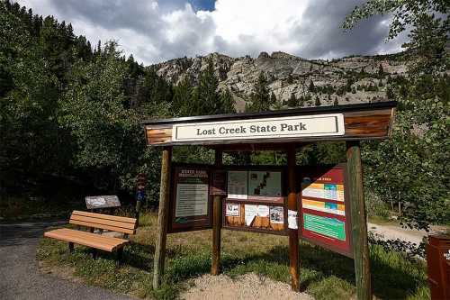 Sign for Lost Creek State Park with a bench nearby, surrounded by trees and mountains under a cloudy sky.