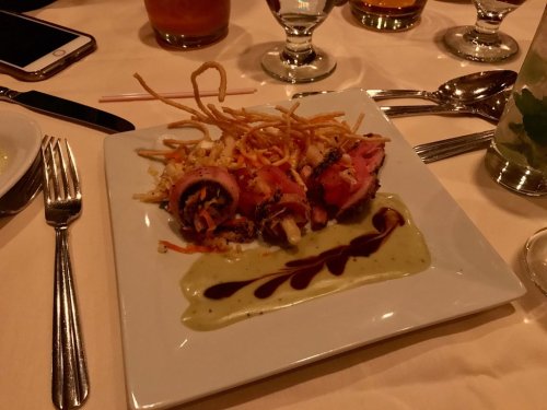 A plated dish featuring sliced meat, crispy noodles, and a drizzle of sauce on a white square plate.