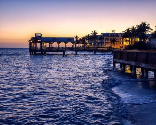 Sunset over a calm ocean with a pier and palm trees, reflecting warm colors in the water.