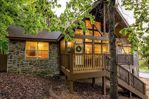 A cozy cabin with stone and wood exterior, surrounded by trees, featuring large windows and a welcoming porch.