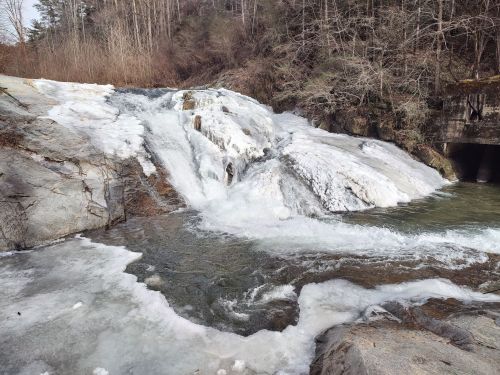 A frozen waterfall cascading over rocky terrain, surrounded by bare trees and icy patches.