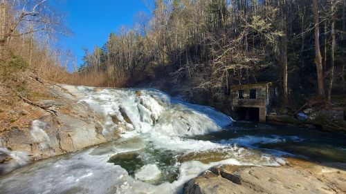 A frozen waterfall flows past a stone building, surrounded by trees under a clear blue sky.