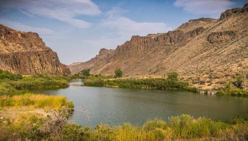 A serene river flows between rugged mountains, surrounded by lush greenery and dry hills under a blue sky.