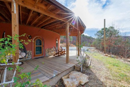 A cozy, round house with a wooden porch, surrounded by nature and sunlight shining through the trees.