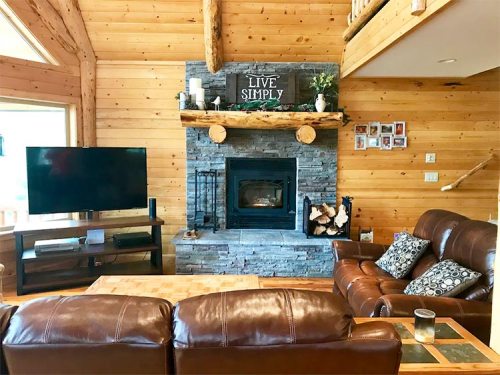 Cozy wooden living room with a stone fireplace, leather sofas, and a "Live Simply" sign above the mantel.