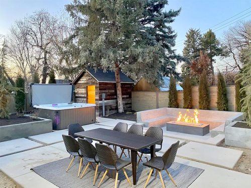 A modern outdoor space featuring a dining table, fire pit, hot tub, and lush greenery at sunset.