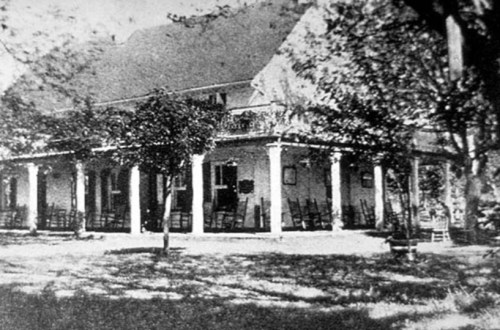 Historic building with a porch supported by columns, surrounded by trees and empty chairs on the lawn.