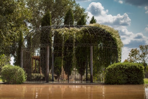 A decorative metal gazebo surrounded by greenery, reflecting in a calm water surface under a blue sky with clouds.