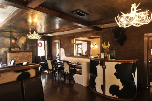 Interior of a rustic restaurant featuring cowhide decor, antler chandeliers, and wooden furnishings.