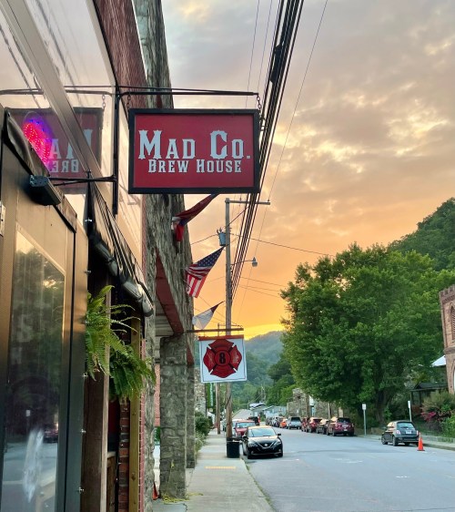 Sign for Mad Co. Brew House at sunset, with trees and a quiet street in the background.
