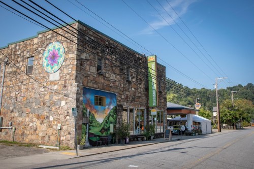 A stone building with colorful murals, a sign, and outdoor seating, set along a quiet street with trees in the background.