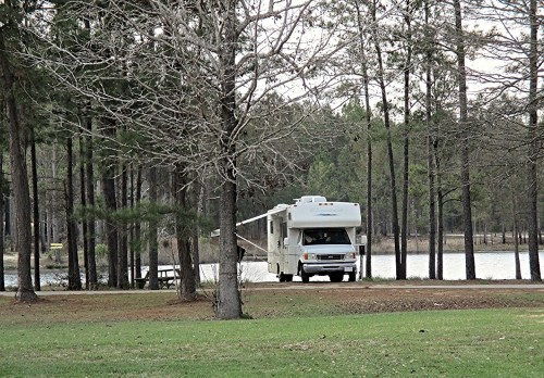 A white RV parked near a lake, surrounded by trees in a serene outdoor setting.