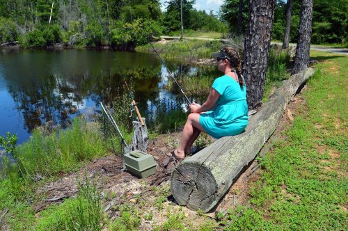 A person in a blue dress sits on a log by a pond, fishing with a rod and reel on a sunny day.