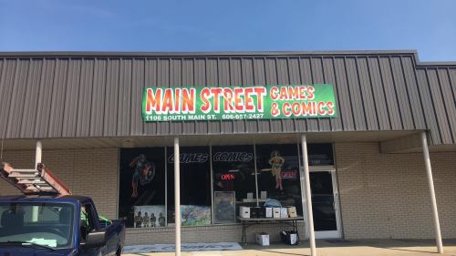 Storefront of "Main Street Games & Comics" with a colorful sign, featuring comic-themed decorations and a blue truck parked outside.