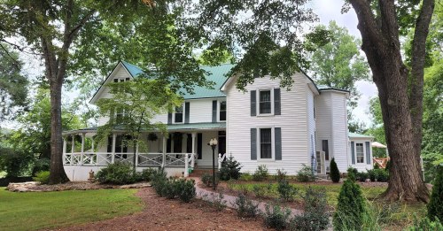 A charming white two-story house with a green metal roof, surrounded by trees and landscaped gardens.