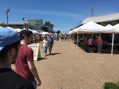 A bustling outdoor market with tents, vendors, and people walking along a paved path under a clear blue sky.