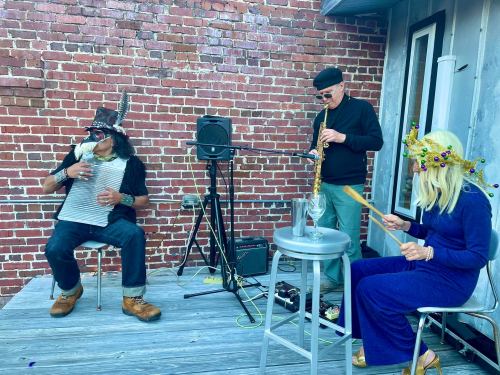 Three musicians perform on a deck against a brick wall, playing an accordion, saxophone, and percussion.
