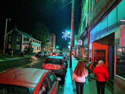 A nighttime street scene with colorful lights, parked cars, and people walking along the sidewalk.