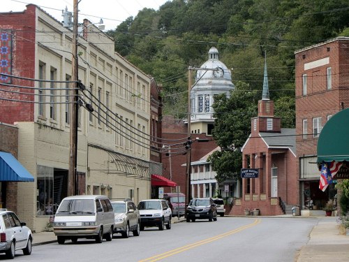 A small town street with brick buildings, a church, and a clock tower, lined with parked cars and power lines.
