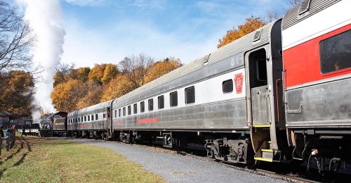 A vintage train with passenger cars parked on a track, surrounded by autumn trees and a blue sky.
