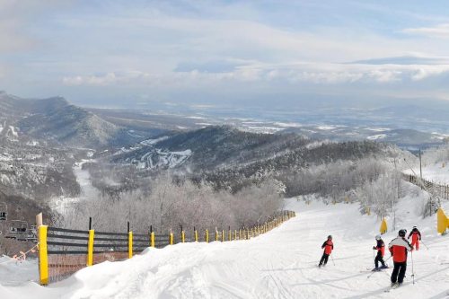 Skiers descend a snowy slope with a scenic view of mountains and valleys under a clear sky.