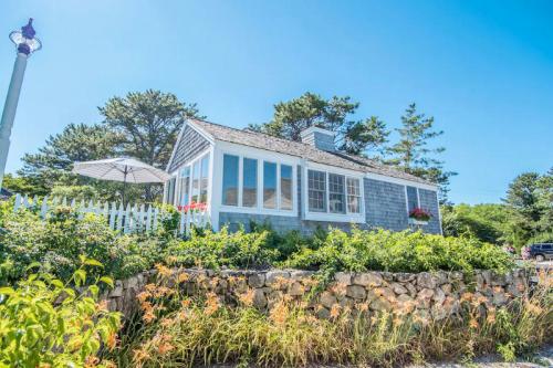 A charming blue cottage surrounded by greenery and flowers, with a white picket fence and an umbrella for shade.