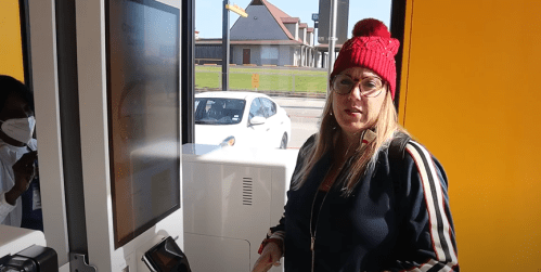 A woman in a red beanie stands at a kiosk, looking at the camera with a serious expression.