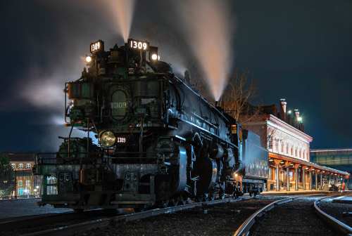 A vintage steam locomotive at night, with smoke billowing and lights shining, parked near a historic train station.
