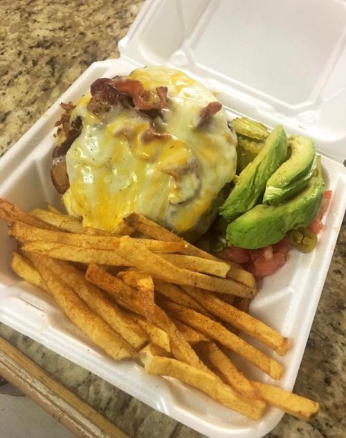 A cheeseburger topped with bacon, served with fries and avocado slices in a takeout container.