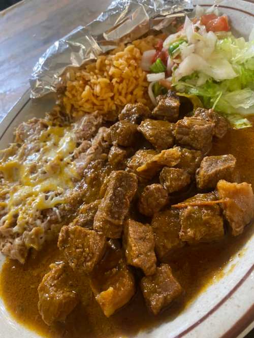 A plate of beef chunks in sauce, served with rice, refried beans topped with cheese, and a side of lettuce and tomato.