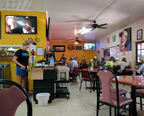 A busy diner with patrons seated at tables, a waitress serving, and TVs showing programs on the walls.