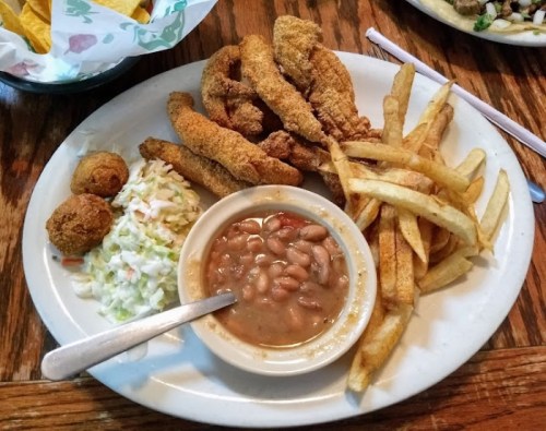 A plate of fried fish fillets, coleslaw, fried okra, baked beans, and French fries on a wooden table.