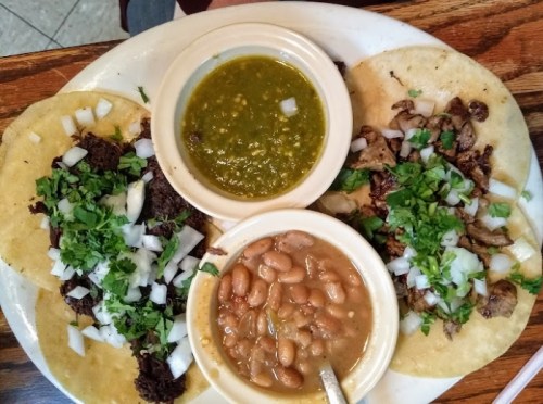 A plate with three tacos topped with onions and cilantro, accompanied by green salsa and a bowl of refried beans.