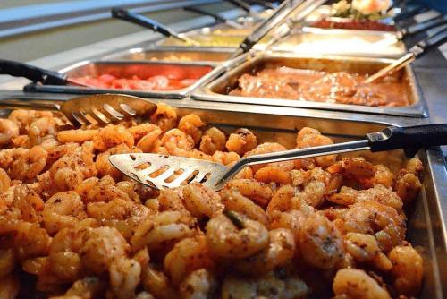 A buffet display featuring trays of seasoned shrimp and various dishes, with serving utensils in the foreground.