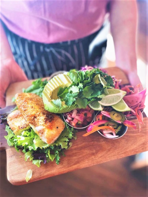 A wooden platter with grilled salmon, fresh greens, lime, and colorful side salads garnished with herbs.